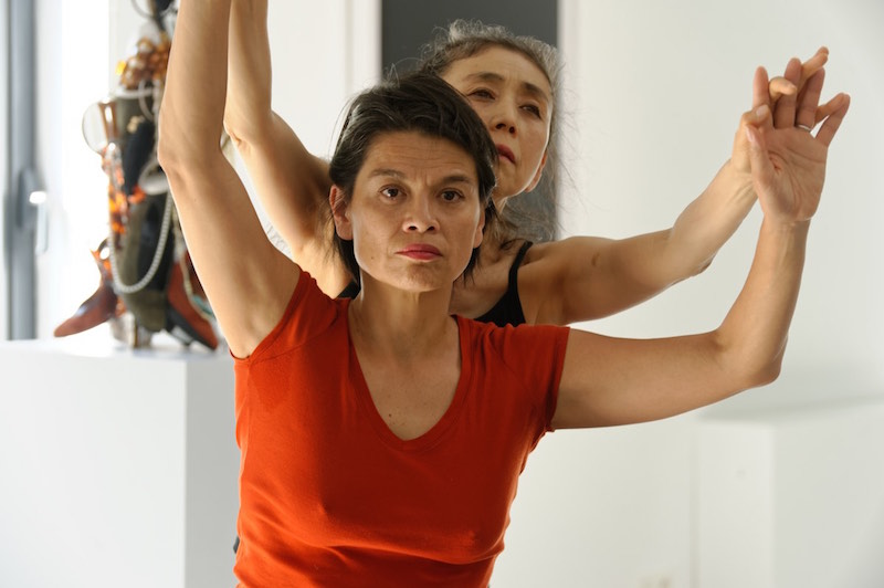 Hunyh in a red t-shirt holds Eiko's fingertips above her head as Eiko stands behind her